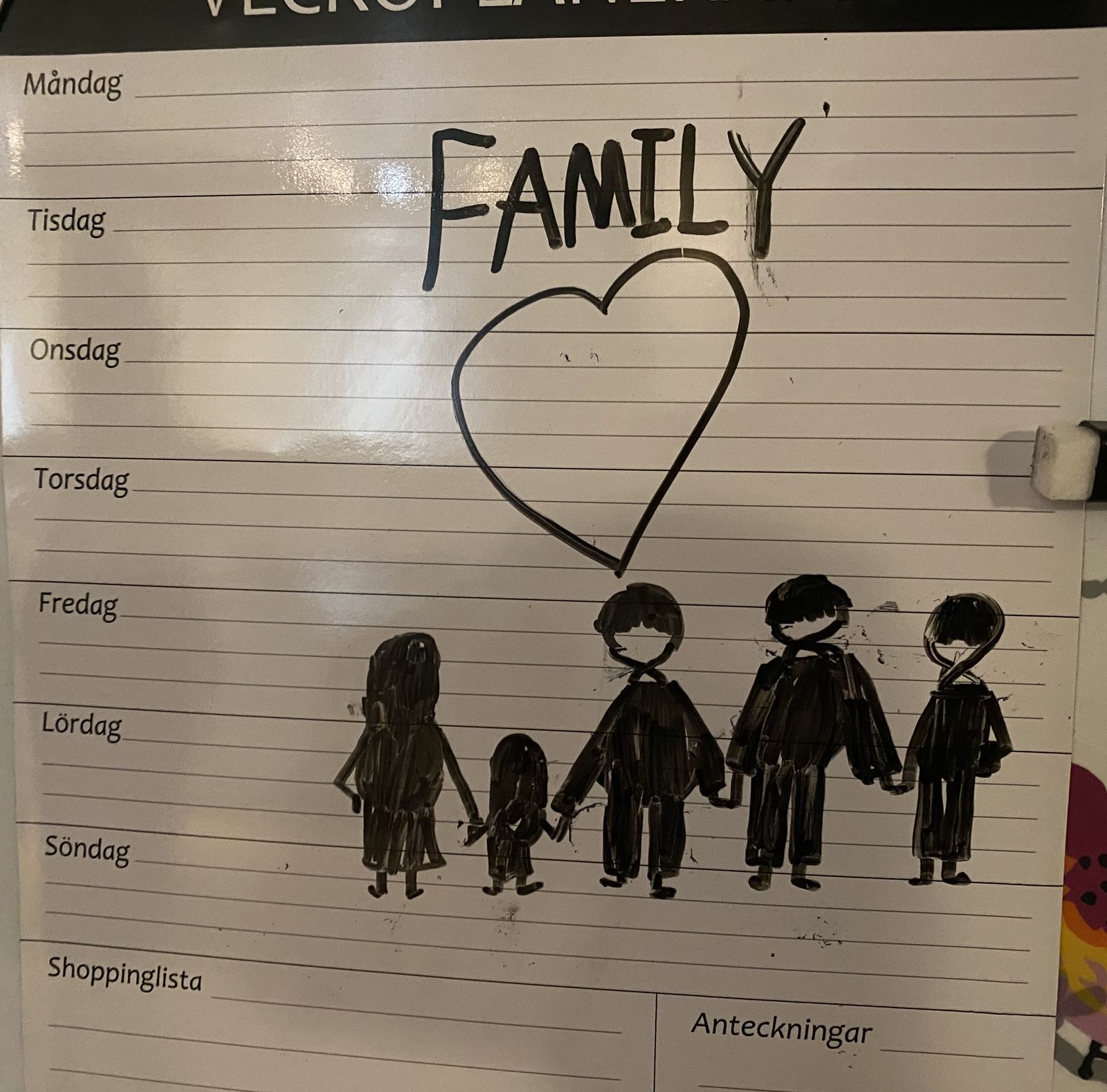 The word "FAMILY" and a heart drawn above five stick figures holding hands, in black marker on a weekly planner on the fridge.