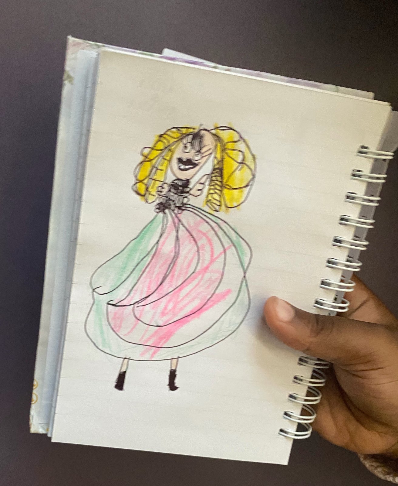 A princess with big yellow hair in a pink and green ball gown, drawn in a spiral notebook held in a hand.