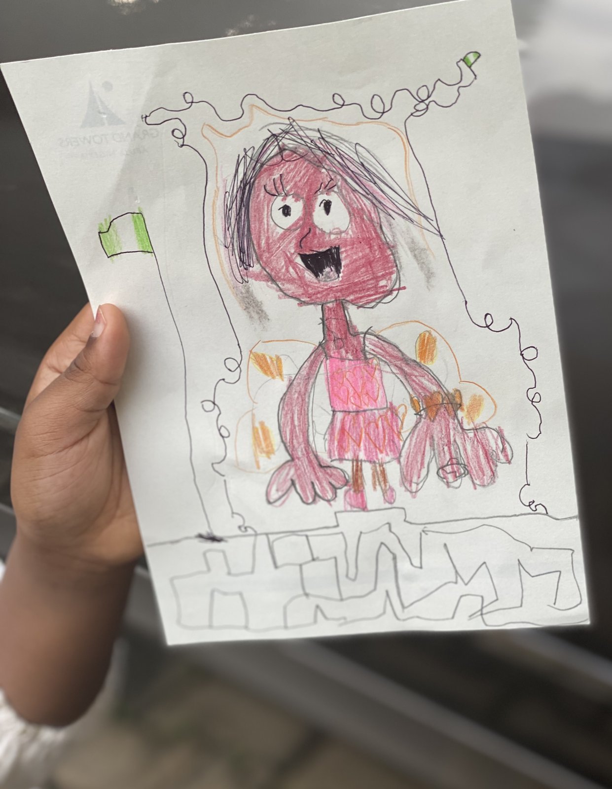 A pink figure with a big smile and wild hair inside a doodled frame, with a small green and white Nigerian flag in the corner. A child's hand holds the paper.